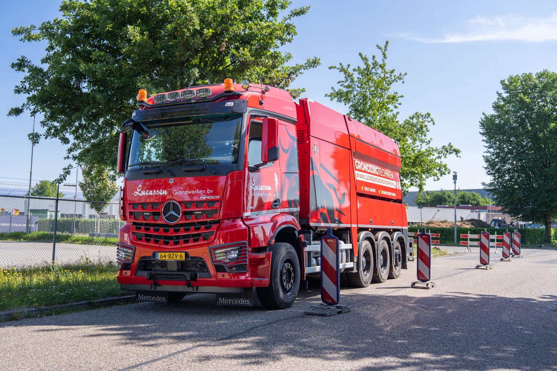 Een rode Mercedes-Benz vrachtwagen van Lucassen Grondwerken B.V. in Overasselt staat geparkeerd langs de weg op een zonnige dag. De vrachtwagen heeft op de zijkant grote zwarte en witte letters en wordt geflankeerd door rood-witte afzethekken.