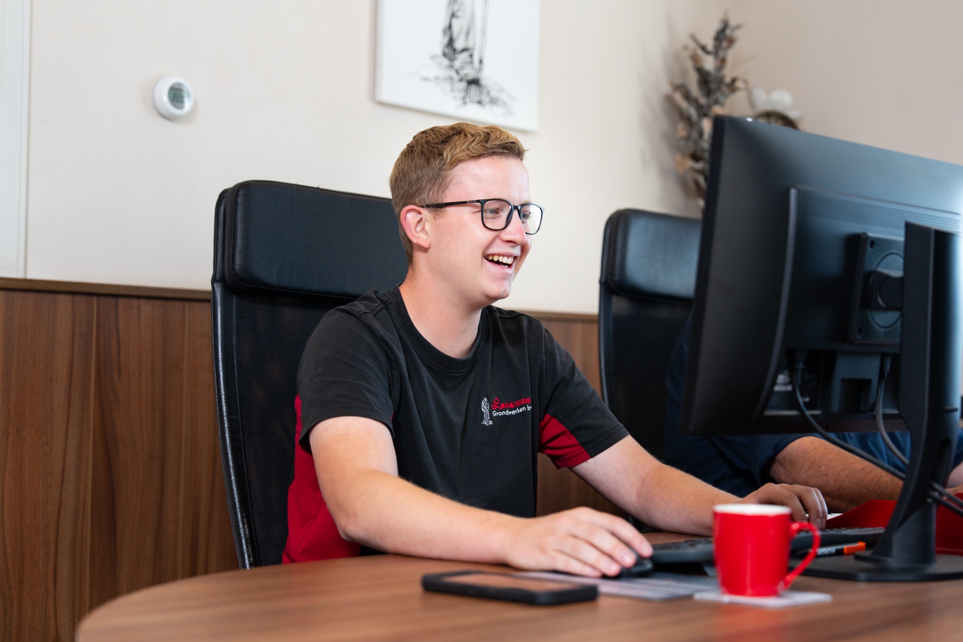 Een lachende, blonde man met een bril en een zwart-rood T-shirt zit achter een bureau in een kantooromgeving en werkt op een desktopcomputer. Hij houdt een muis vast en kijkt naar het scherm. Er staat een rode mok op het bureau.