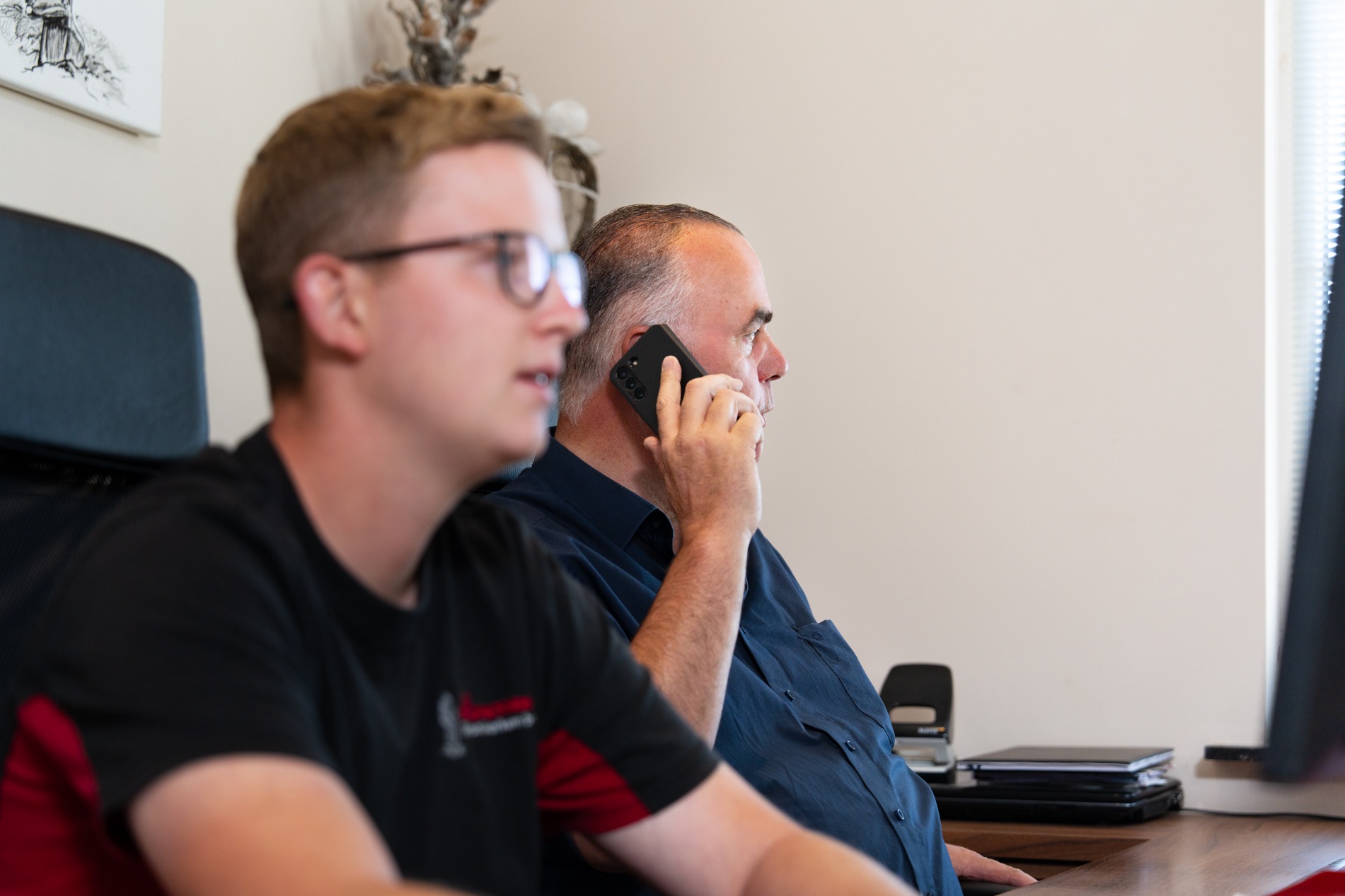 Twee mannen zitten naast elkaar achter een bureau. De man op de voorgrond is jonger, draagt een bril en een zwart-rood T-shirt, en kijkt naar een computerscherm (buiten beeld). De oudere man rechts zit in profiel, draagt een donkerblauw overhemd en is aan het bellen met een zwarte smartphone.