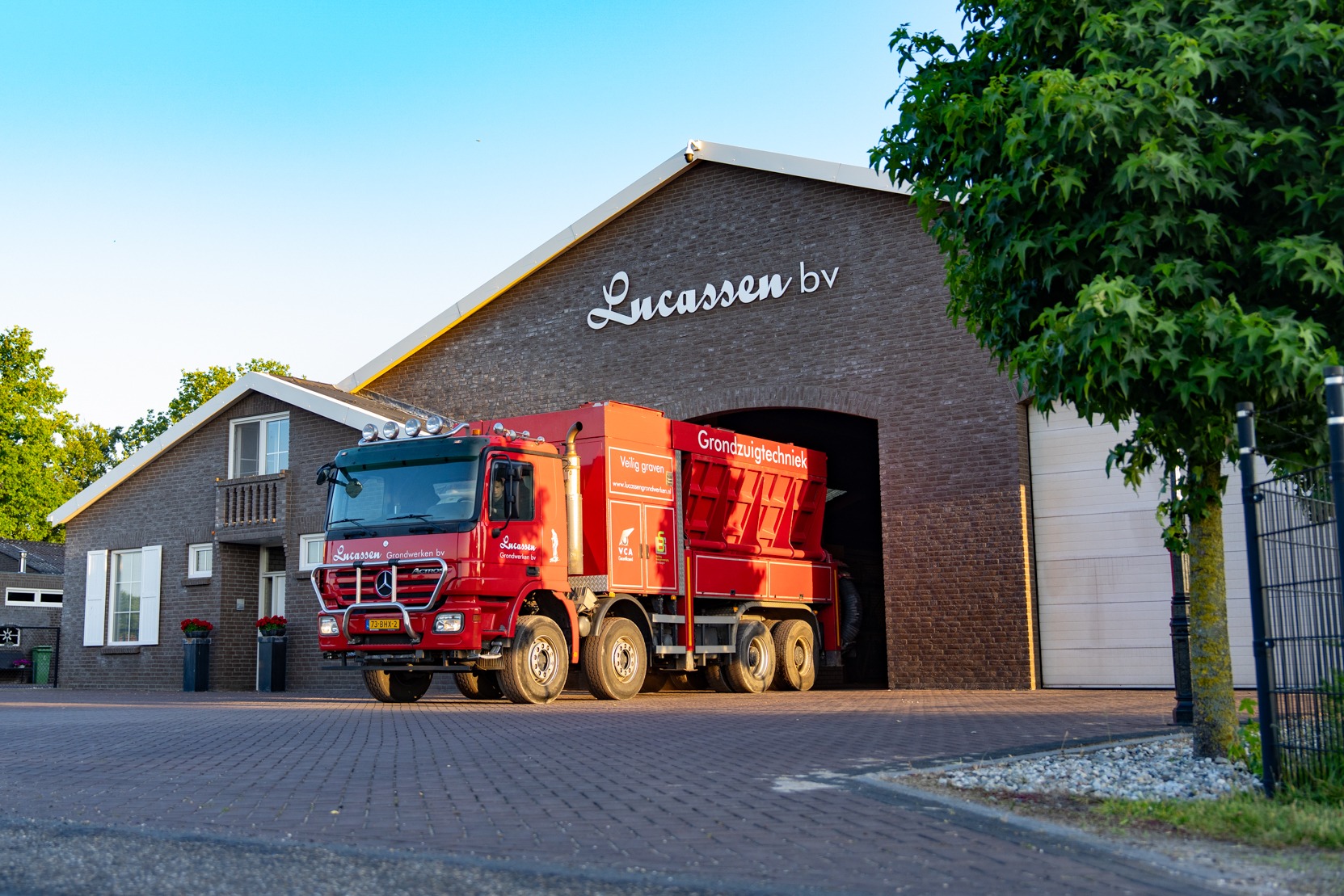 Een grote, rode vrachtwagen van Lucassen Grondwerktechniek staat voor een bakstenen bedrijfsgebouw met de witte letters "Lucassen bv" boven de garagepoort. Het is een zonnige dag en er staat een boom rechts in beeld.