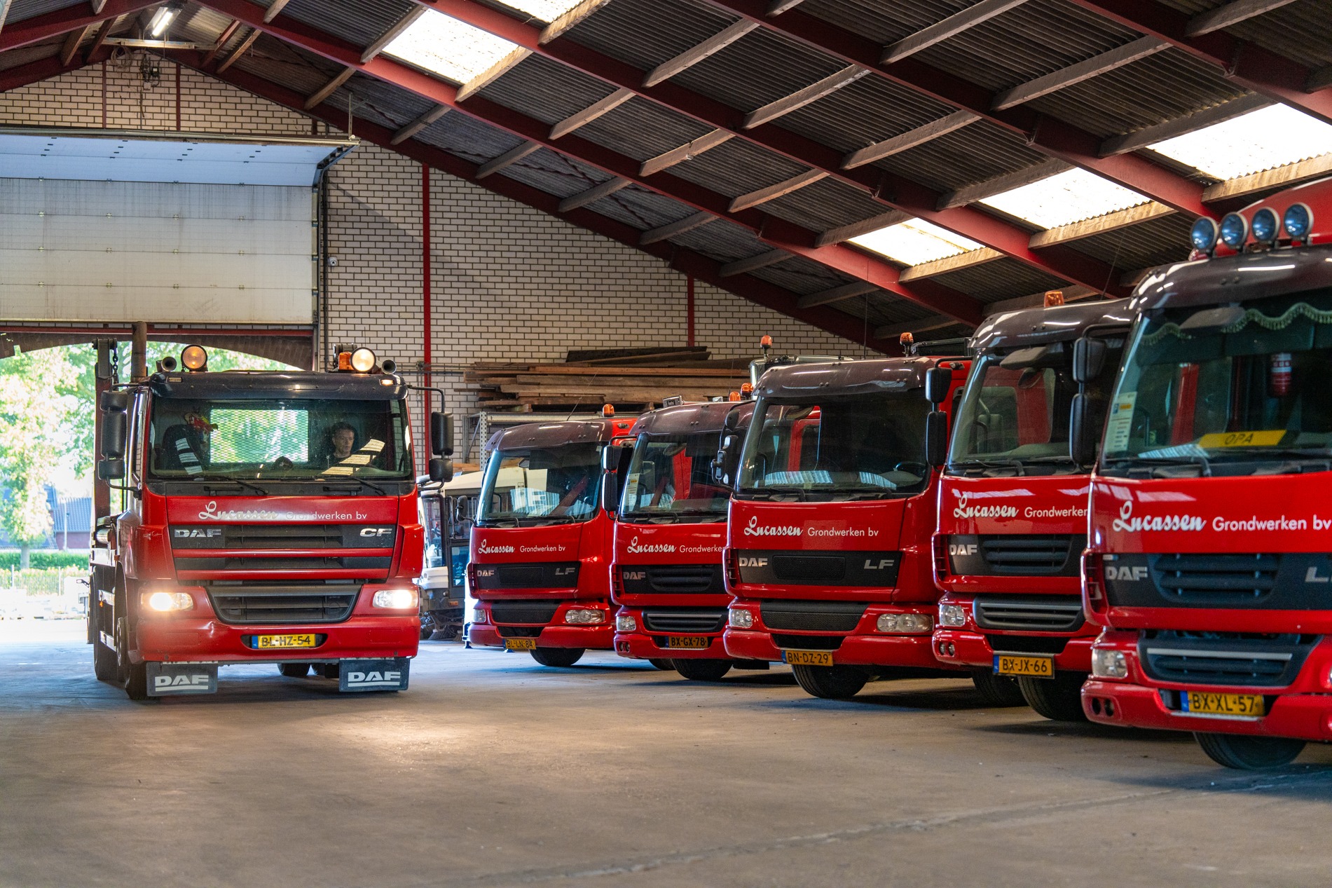 Een rij rode DAF-vrachtwagens met de branding van Lucassen Grondwerken bv staat geparkeerd in een ruime opslagloods met een bakstenen achterwand en een metalen dakconstructie. De voorste vrachtwagen (links) rijdt langzaam de loods in of uit.