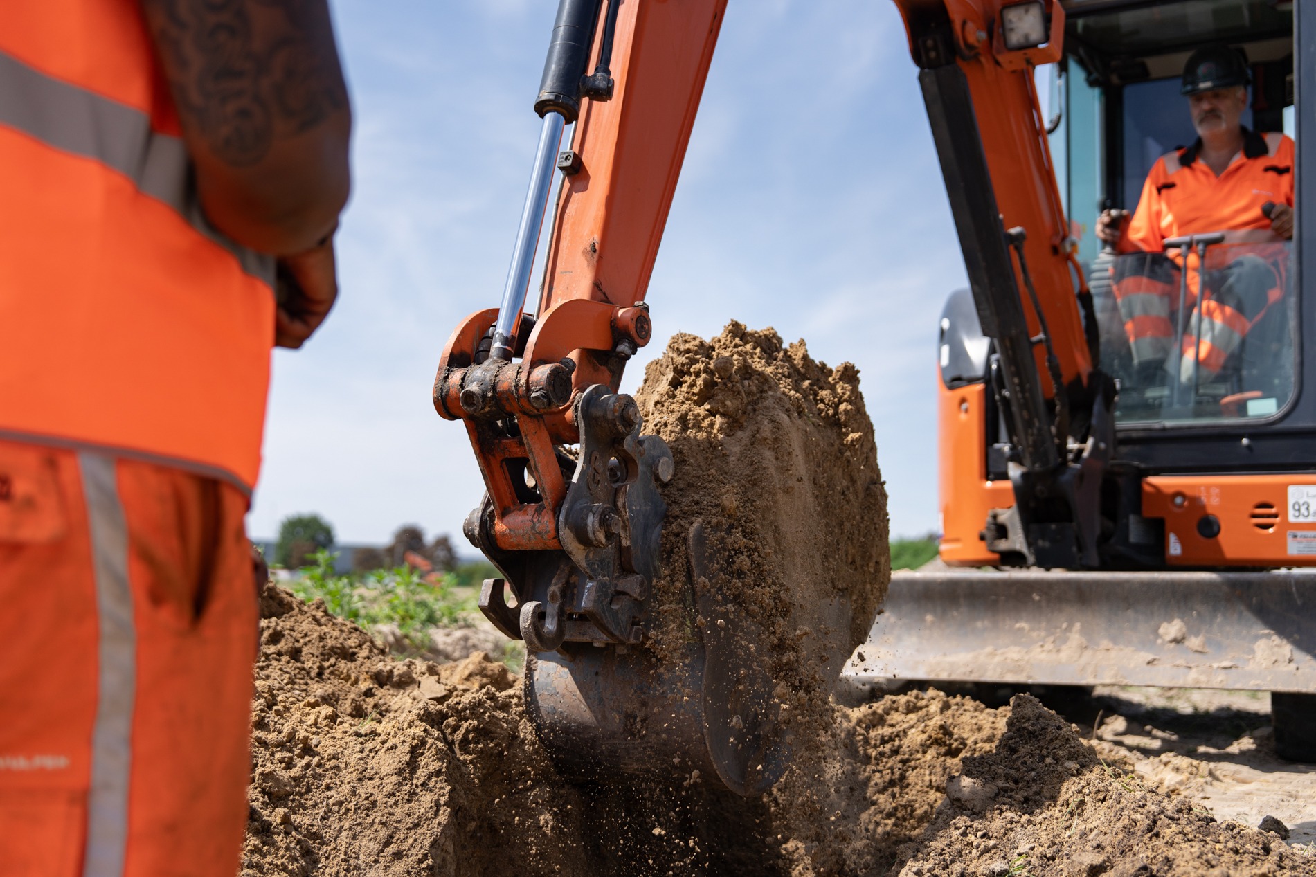 Een close-up van de bak van een oranje mini-graafmachine in Overasselt die bezig is met grondwerk, scheppend in een hoop aarde. In de cabine zit de bestuurder, gekleed in oranje werkkleding. Op de voorgrond, met de rug naar de camera, staat een persoon in oranje werkkleding die de werkzaamheden overziet.